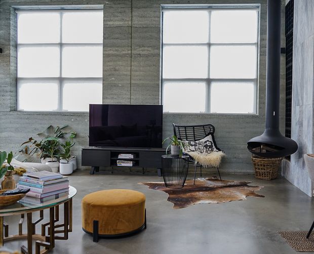 Industrial-style living room with polished concrete flooring, exposed textured concrete walls, large divided-panel windows, a black suspended wood-burning fireplace, low black TV console, television, wire lounge chair, leather ottoman, nesting glass tables, indoor plants, and layered neutral furnishings. The space has a high ceiling and a strong mix of concrete, black metal, timber, leather, and warm ochre accents.