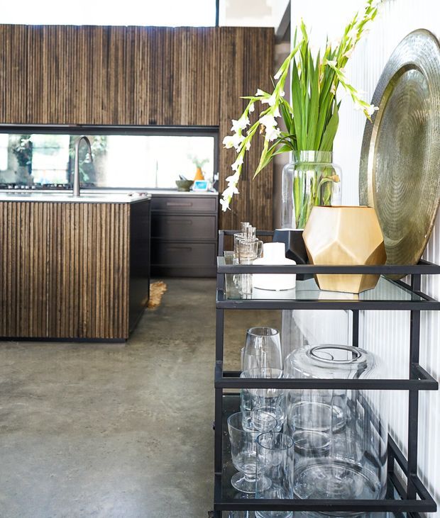 Bright contemporary kitchen and entertaining area with dark vertically fluted timber cabinetry, a concrete floor, stainless-steel sink, and black-framed glass shelving displaying glassware. A large metallic circular wall feature, geometric ceramic decor, and tall leafy floral arrangement add visual interest.