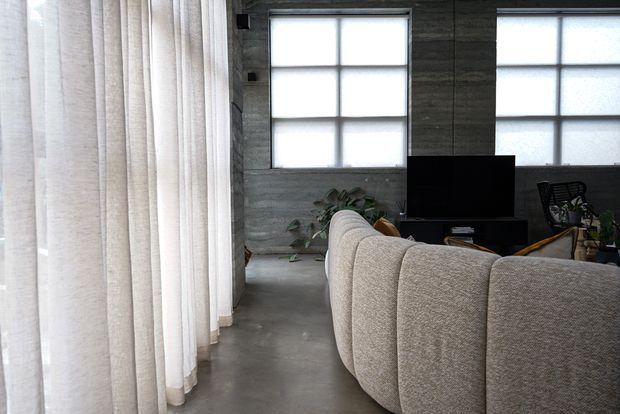 A contemporary living room with a large curved textured sectional sofa facing a television. The space features polished-looking concrete flooring, dark grey concrete or plaster walls, frosted gridded windows, sheer floor-to-ceiling curtains, and sparse indoor planting. A desk and chair are visible in the background.