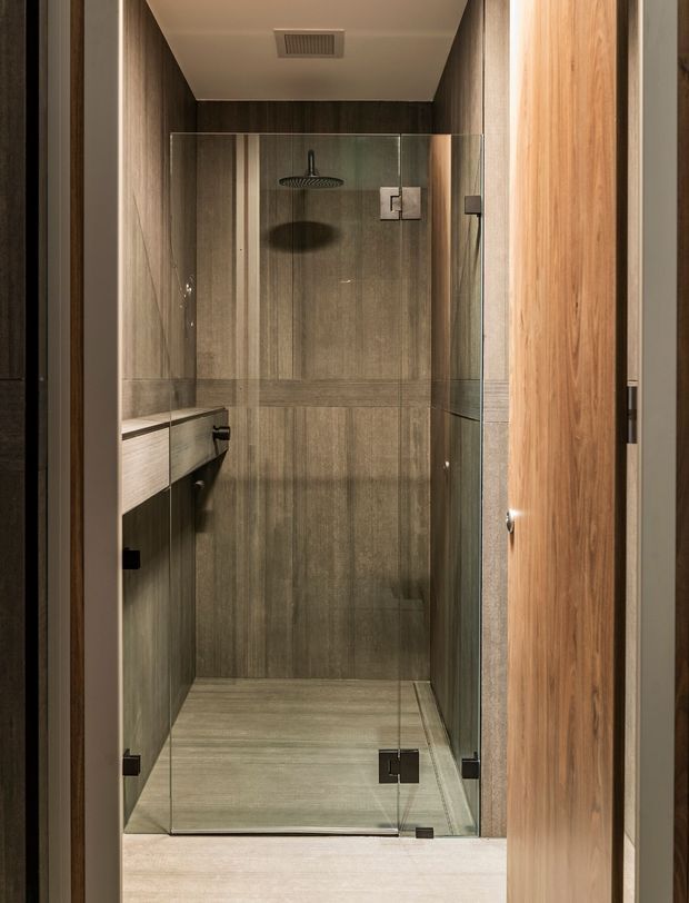 A compact contemporary bathroom shower enclosure viewed through a timber-framed doorway. The walk-in shower has clear frameless glass panels and dark hardware, with large vertically grained grey-brown stone-look tiles covering the walls and floor. A floating timber-toned vanity or shelf is visible along the left wall, while warm timber joinery frames the entrance. Recessed ceiling ventilation and a rainfall shower complete the understated, warm-toned design.