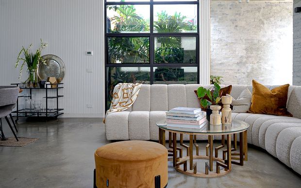 A stylish open-plan living room with a large cream modular sectional sofa, round glass coffee table with sculptural timber base, and a mustard velvet ottoman. The room features a polished concrete floor, textured light stone or masonry feature wall, vertical white wall panelling, black-framed floor-to-ceiling glazing, and contemporary decorative styling with indoor plants and neutral-toned accessories.