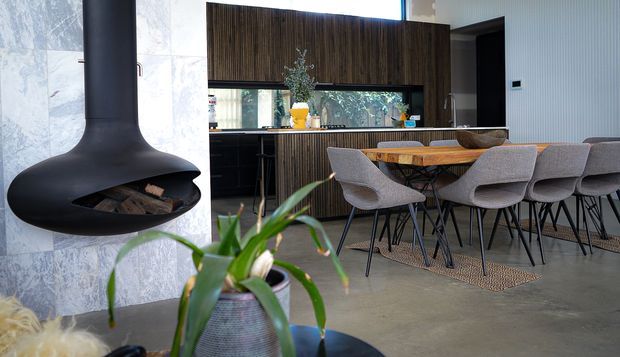 A contemporary open-plan kitchen and dining area with dark vertical timber cabinetry, a long island bench, and a timber dining table surrounded by upholstered dining chairs. A sculptural black suspended fireplace is mounted against a pale stone feature wall, with polished concrete flooring and large windows bringing in natural light.