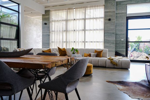 Bright contemporary open-plan living and dining area with floor-to-ceiling glazing, sheer curtains, polished concrete flooring, textured grey feature walls, a large sectional sofa, and a substantial live-edge timber dining table with upholstered chairs. The room has a spacious, airy feel and strong indoor-outdoor connection.