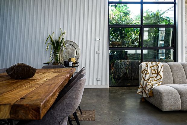 Industrial-style dining and living area with a substantial live-edge timber dining table, upholstered dining chairs, a curved textured sofa, ribbed wall panelling, polished concrete flooring, and large black-framed industrial glazing overlooking greenery. Neutral furnishings and sculptural decor create a refined, contemporary atmosphere.