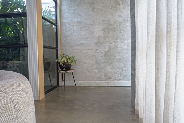 A softly lit contemporary interior corner with a pale exposed brick feature wall, polished concrete floor, black-framed glazed doors or windows, and full-height sheer white curtains. A small plant stand and the edge of a textured upholstered sofa are visible, with greenery seen through the glazing.
