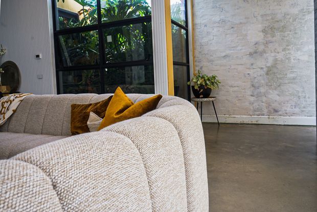 A contemporary living room corner featuring a large textured cream sectional sofa with mustard and brown cushions. The space has a polished concrete floor, a pale textured masonry feature wall, dark-framed floor-to-ceiling glazing, and vertical wall panelling, creating a warm industrial-contemporary atmosphere.