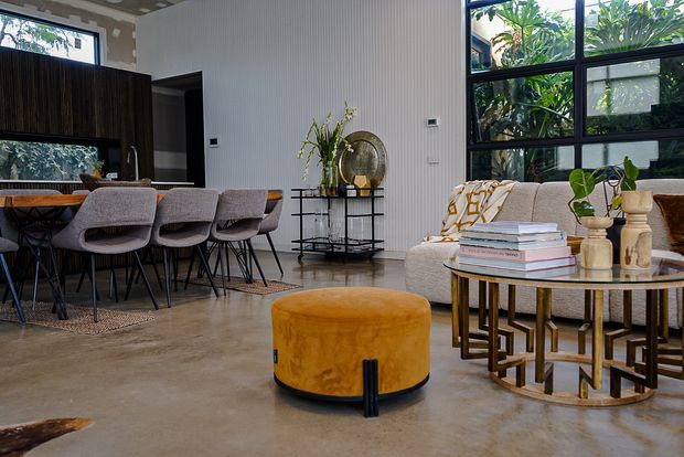 Open-plan contemporary living and dining area with polished concrete flooring, a dark timber kitchen or dining screen, a long dining table with upholstered chairs, and a lounge seating area beside expansive black-framed glazing. Vertical wall panelling, sculptural decor, a round mustard ottoman, and nested brass-toned coffee tables add warmth and contrast.