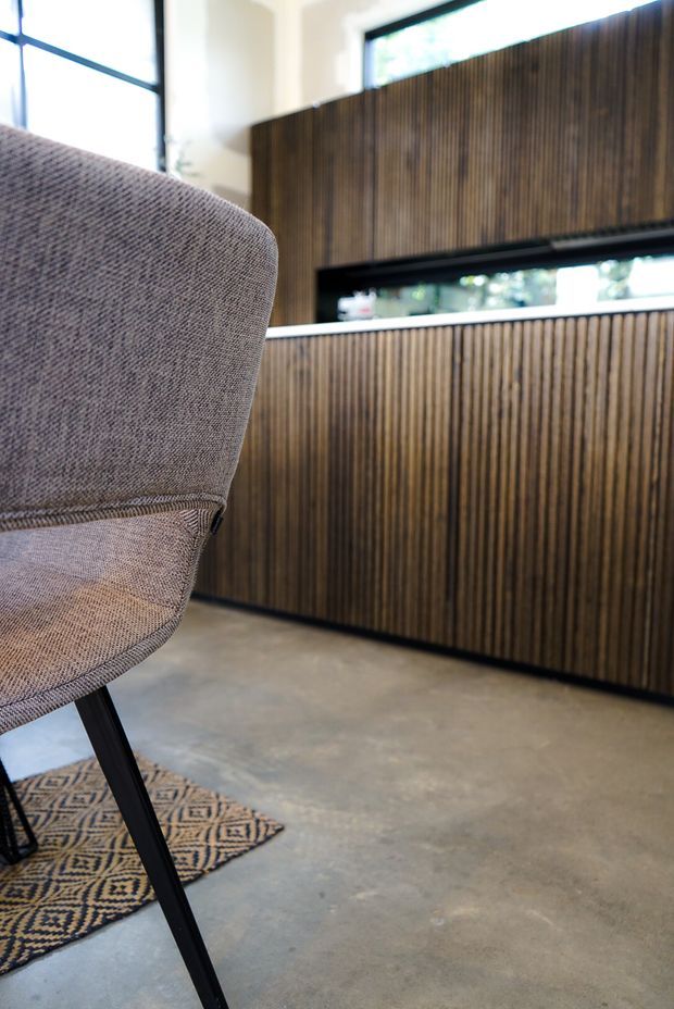 Close-up interior view of a refined contemporary space featuring a textured upholstered armchair in the foreground, dark timber vertical slatted built-in cabinetry or wall panelling, slim black-framed horizontal glazing, and a polished concrete floor. A patterned area rug adds a subtle decorative accent.