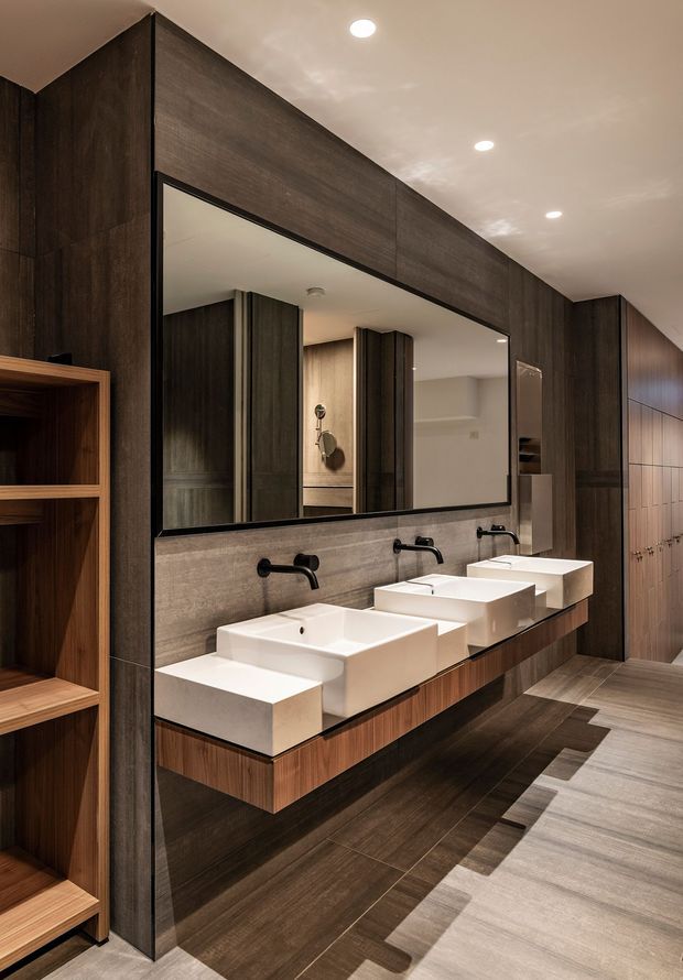 A refined contemporary bathroom featuring a long double vanity with multiple white vessel-style rectangular basins, matte black wall-mounted tapware, dark timber-look wall cabinetry, warm timber vanity joinery, large-format dark floor tiles, and a full-width mirror. Open shelving is visible at the left, while a partially reflected adjoining vanity or bathroom area appears in the mirror.