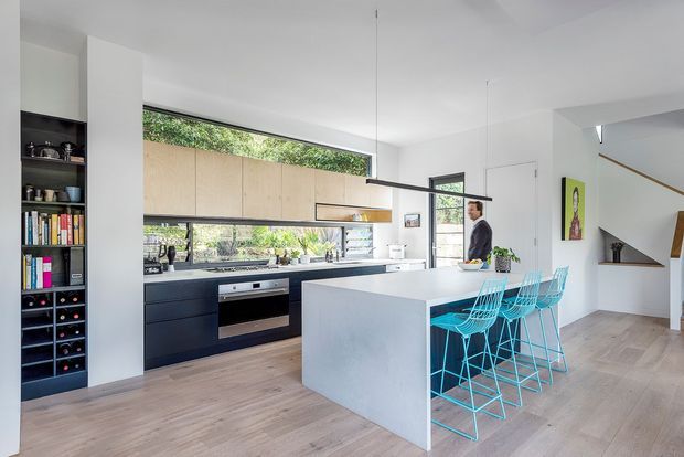 Bright contemporary open-plan kitchen with a large white island and vivid turquoise bar stools. The room features dark lower cabinetry, light timber overhead cabinetry, integrated appliances, open shelving, a broad horizontal window, pale timber flooring, and a minimalist linear pendant light. A stair and adjoining living area are visible in the background.