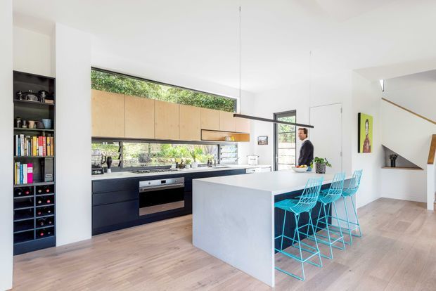 Bright contemporary open-plan kitchen and dining area with a long dark lower kitchen run, light timber upper cabinetry, white island bench, and vivid turquoise wire bar stools. Full-height glazing brings in greenery, while an open stair and built-in shelving are visible at the right and left edges. Pale timber flooring, white walls, and a slim linear pendant create an airy, clean interior.