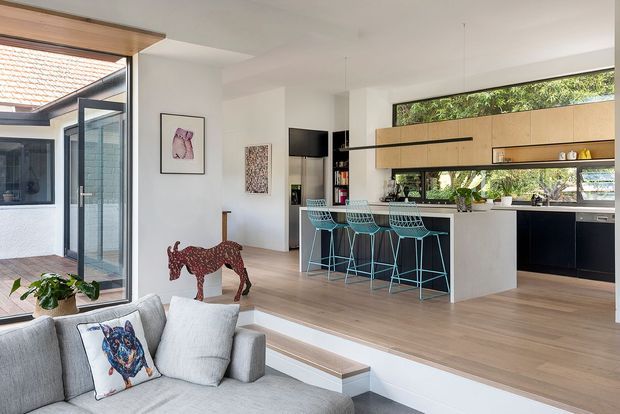 Open-plan contemporary kitchen and living area with a raised timber-finished floor platform, pale cabinetry, a large island with turquoise wire bar stools, and extensive horizontal glazing overlooking greenery. A grey sofa occupies the foreground, with a glazed indoor-outdoor connection at left, light timber ceiling detailing, pendant lighting, and playful artwork.