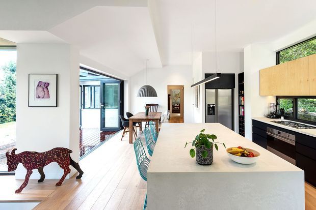 Bright open-plan kitchen and dining area with a long pale island, dark lower cabinetry, light timber upper cabinets, stainless-steel appliances, and extensive glazing. A dining table sits behind the island, with a living area and outdoor greenery visible through the glass. The space has clean contemporary lines, a light neutral palette, and abundant natural light.