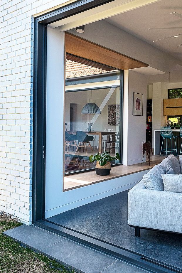 A bright contemporary open-plan interior viewed through a wide dark-framed glazed opening. The foreground shows a light grey sectional sofa on dark carpet, while the adjoining dining area is visible through the glazing with timber furniture, a sculptural pendant light, and indoor planting. White-painted brickwork forms the exterior wall, and a timber-lined structural opening frames the transition between spaces. A compact kitchen with a timber island and turquoise bar stools is visible in the background.