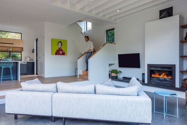 Bright open-plan living room with a large pale sectional sofa in the foreground, a wall-mounted television, and a built-in fireplace with a lit fire. The room connects to a kitchen and dining area, with a timber staircase leading to an upper level. White walls, light timber flooring, exposed white ceiling beams, and contemporary built-in shelving create an airy, refined interior.