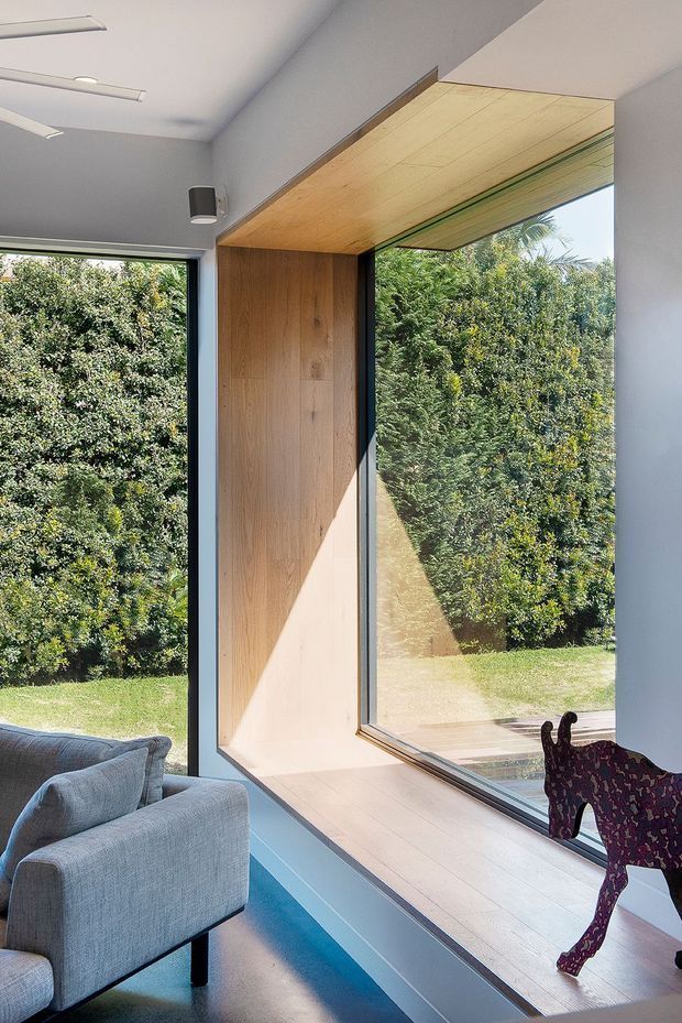 Bright contemporary interior corner with floor-to-ceiling glazing overlooking a leafy garden. A grey upholstered sofa sits beside a deep window seat or built-in timber-lined alcove, with a warm light timber ceiling and wall lining, slim dark window frames, and a playful sculptural animal figure in the foreground.