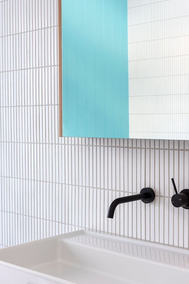 Close-up of a contemporary bathroom vanity area featuring a white bathtub or basin, vertically arranged narrow white wall tiles with light grout, and matte black wall-mounted tapware. A large turquoise mirror or reflective panel with a slim timber-toned frame adds a vivid colour accent.