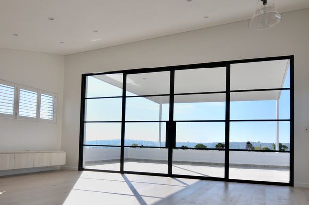 Bright, mostly empty contemporary interior with pale timber flooring, white walls and ceiling, black-framed floor-to-ceiling glazed doors or windows opening to a balcony or terrace with expansive water and landscape views. White plantation shutters and a simple clear pendant light are visible, with strong geometric shadows cast across the floor.