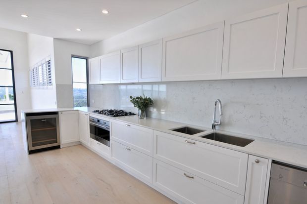 Bright, spacious white kitchen with shaker-style cabinetry, a long stone benchtop and matching full-height splashback, integrated gas cooktop and oven, double undermount sink, and stainless-steel tap and dishwasher. Light timber flooring, recessed ceiling lights, and large windows create an airy, refined interior.