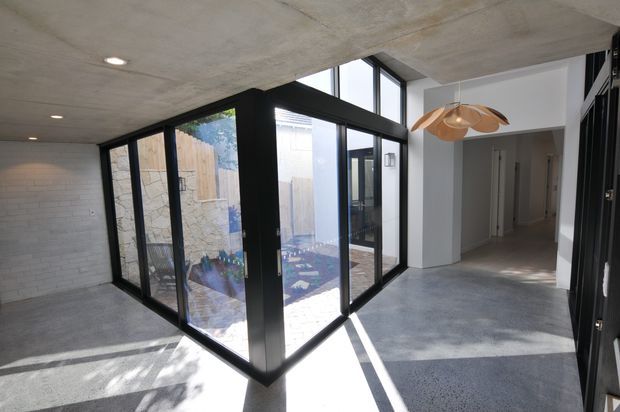 Contemporary concrete interior with polished concrete flooring, exposed concrete ceiling, black-framed floor-to-ceiling glazing, and a glazed corner opening onto a small courtyard. A timber sculptural pendant hangs near a white hallway and circulation area.