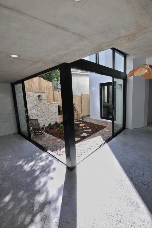 Interior view of a contemporary courtyard-facing space with large black-framed floor-to-ceiling glazing, exposed concrete ceiling, polished concrete floor, and a central structural column. The glazing opens visually onto a small landscaped courtyard with brick paving, stone walling, timber fencing, and planted garden beds. A sculptural pendant light is visible at the upper right.
