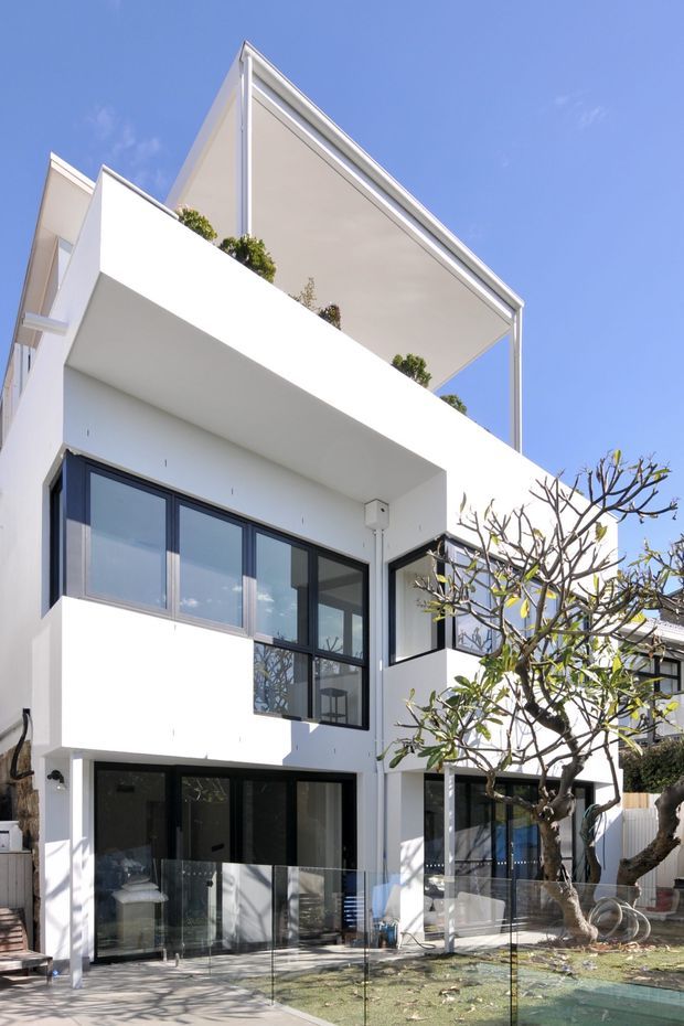 A bright contemporary multi-storey residential facade with crisp white rendered walls, extensive dark-framed glazing, projecting balconies, and a dramatic pitched roof canopy. A transparent glass balustrade encloses the lower outdoor terrace, while mature ornamental planting sits around the building. The image is photographed from outside under a clear blue sky.