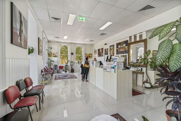 Bright reception area in a clinic or professional practice, featuring a white reception counter, brochure displays, artwork, tiled flooring, waiting chairs, indoor plants, and large windows. The space has a clean, welcoming commercial fit-out with a suspended ceiling and recessed fluorescent lighting.