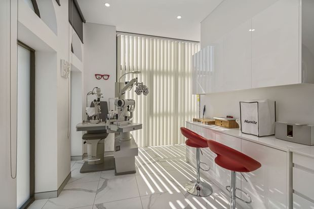 Bright, minimalist optometry consultation and eyewear workspace with white glossy cabinetry, a streamlined counter, two red bar stools, an examination instrument, marble-look tiled flooring, and strong striped daylight from vertical blinds. The room has a clean contemporary clinical-commercial fit-out.