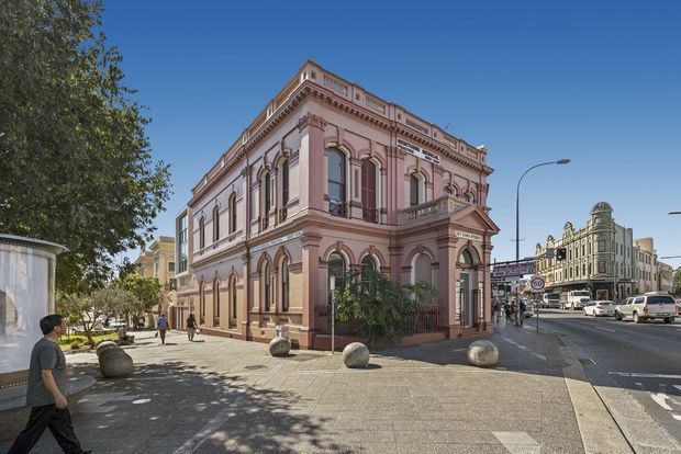 Street-level view of an ornate historic two-storey corner building with a pink-painted masonry facade, arched windows, pilasters, elaborate cornices, decorative parapets and a prominent pediment. It fronts a broad urban sidewalk and intersection, with other heritage-style commercial buildings visible along the street.