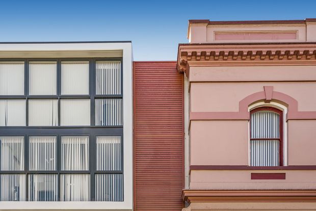 Street-facing view of two adjoining urban building facades: a contemporary white-framed glazed facade with vertical blinds on the left, and an ornate pink-toned heritage-style facade with decorative cornice, arched window surround, and horizontal cladding between the buildings. Clear blue sky above.