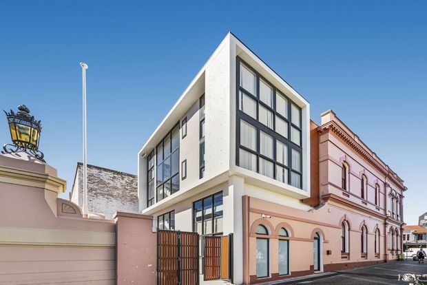 Street-level exterior view of a contemporary multi-storey building inserted between ornate historic masonry buildings. The main building has a sharp projecting upper volume, white rendered or concrete surfaces, extensive black-framed floor-to-ceiling glazing, and a gated ground-level frontage. Adjacent heritage buildings feature pink-toned rendered facades, arched windows, decorative cornices, and traditional detailing.