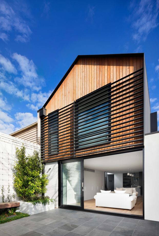 Modern two-storey residential facade viewed from a paved courtyard. The upper level features warm vertical timber cladding screened by prominent black horizontal louvres, with glazing behind. The ground floor has a large open glazed living-room frontage, white rendered walls, dark-framed sliding doors, and a grey stone-paved outdoor area. The building has a strong gabled roof form and a clean, contemporary composition beneath a bright blue sky.