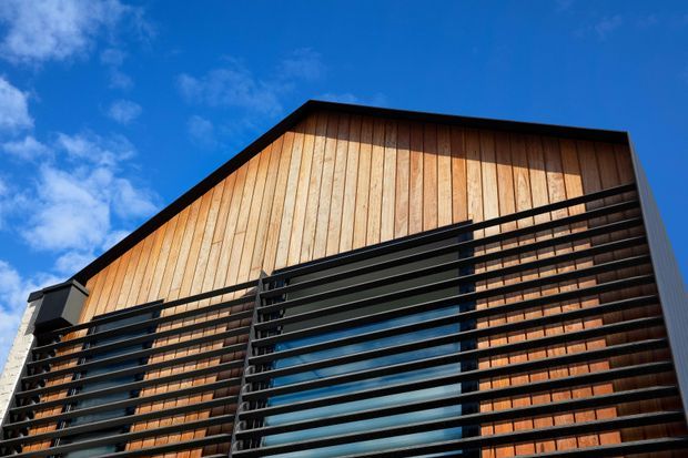 Upward-looking exterior view of a modern multi-storey building facade. The elevation features warm vertical timber cladding, extensive dark horizontal sun-shading or balcony rails, large blue-tinted windows, and a steep asymmetrical gable-like roofline against a clear sky.