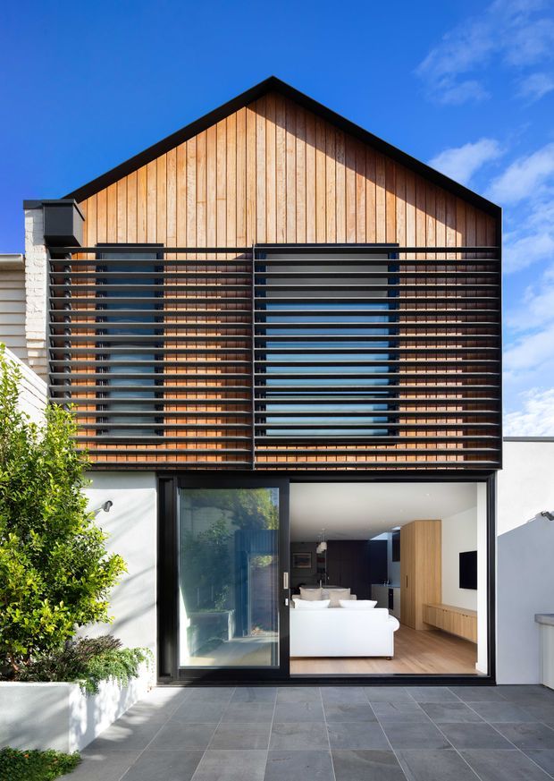 Front exterior view of a contemporary multi-level residence. The upper storey features warm vertical timber cladding beneath a dark gabled roof, with extensive horizontal dark screening over windows and balconies. The ground level has black-framed glazing opening to a tiled terrace, with a minimalist living room visible inside. White rendered walls, landscaped planters, and grey paving complete the facade.