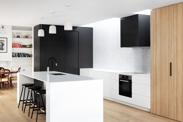 Bright contemporary kitchen with a large white island, integrated sink and black tap, three black bar stools, matte black full-height cabinetry, white lower cabinetry, a speckled light backsplash, and light timber flooring. A timber pantry or tall storage wall occupies the right side, while an adjacent dining area is visible to the left.