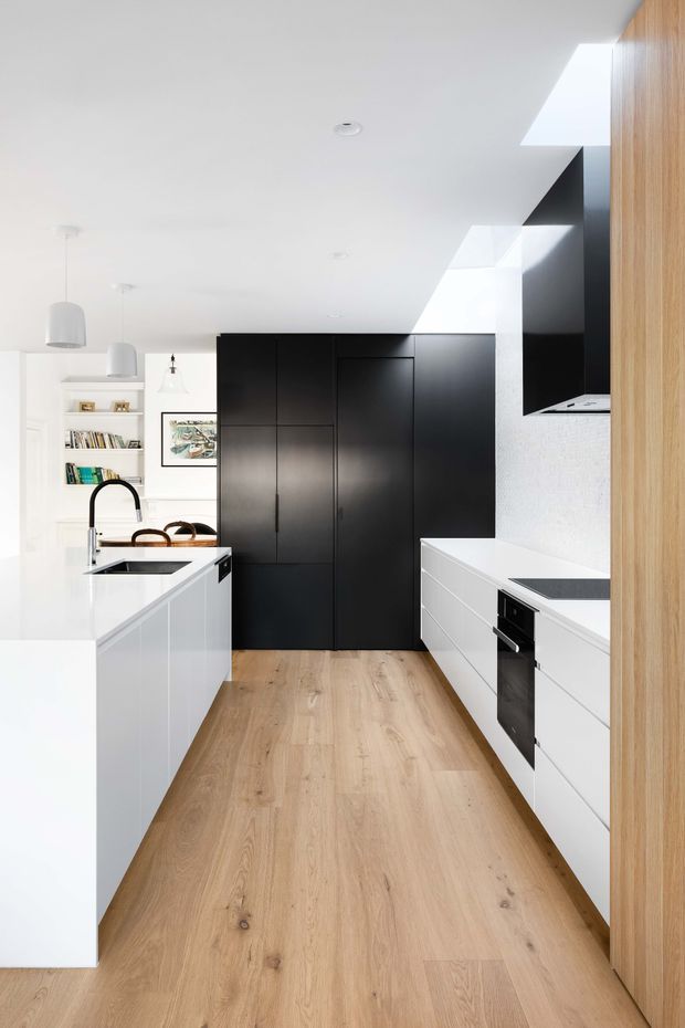 A bright, contemporary galley-style kitchen with a central white island, integrated sink and curved black tap, white cabinetry, pale timber flooring, and a dramatic wall of matte-black full-height joinery. A white benchtop and dark cooktop run along the right side beneath a black-and-white rangehood surround. Skylights or roof glazing introduce strong natural light, while open shelving and artwork are visible at the rear.