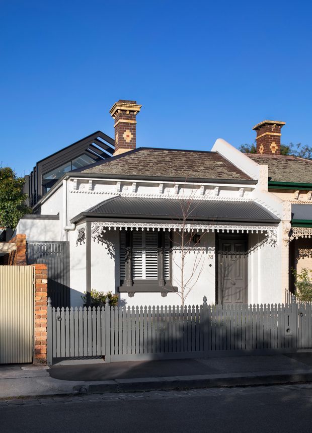 Street-facing view of a compact heritage terrace or cottage-style residence with a white rendered facade, ornate lacework veranda, dark corrugated metal awning, timber picket fence, brick chimneys, and a contemporary glazed roof extension visible behind the front roofline. The facade is strongly Victorian-era in character, with decorative brackets and fretwork.