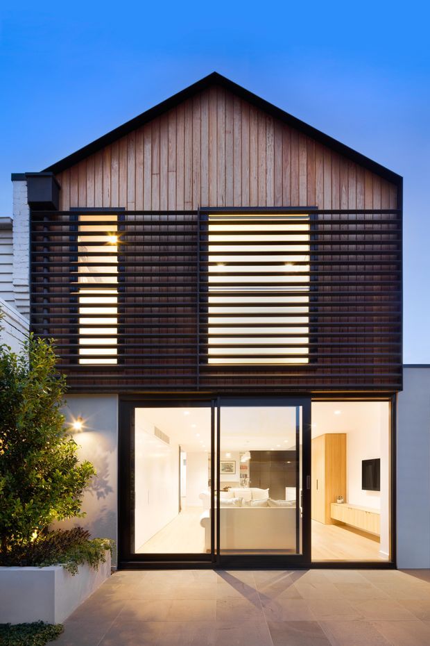 Front exterior view of a contemporary two-storey residence at dusk. The upper gable facade is clad in vertical timber boards and screened by a dark horizontal slatted sun-shading structure, with warm interior light visible through large glazed doors and windows below. A paved entry terrace, rendered walls, and landscaped planter frame the facade.