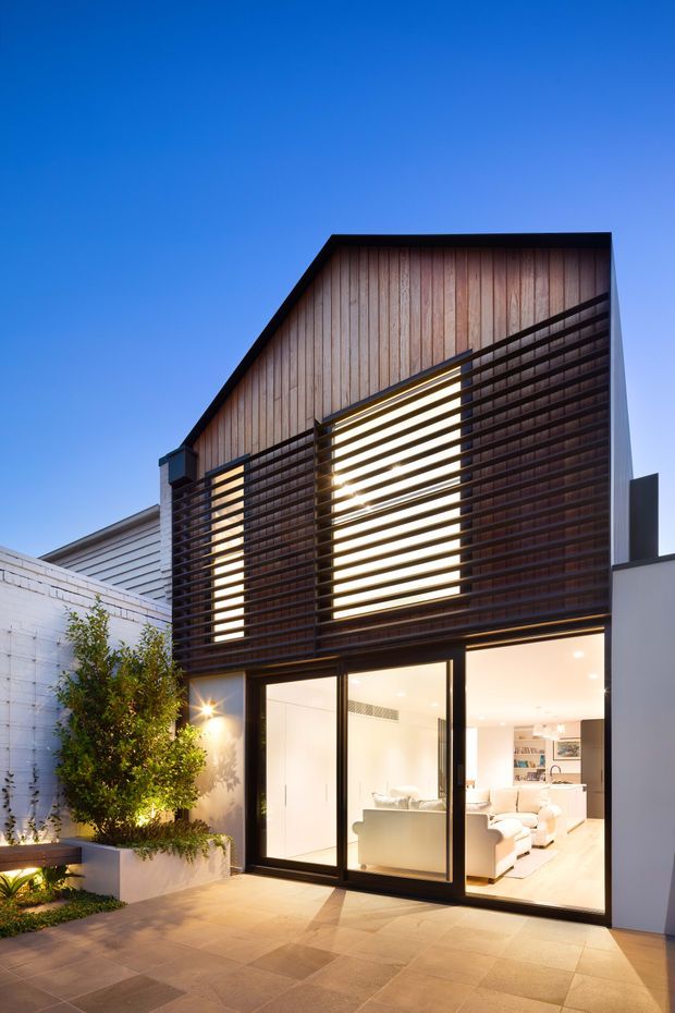 Exterior dusk view of a narrow contemporary multi-storey residence. The upper facade combines vertical timber cladding with dark horizontal louvres screening large windows, while the ground floor has extensive dark-framed glazing opening onto a paved terrace. Warm interior lighting and a visible living room appear through the glazing, with a small landscaped courtyard along the side.