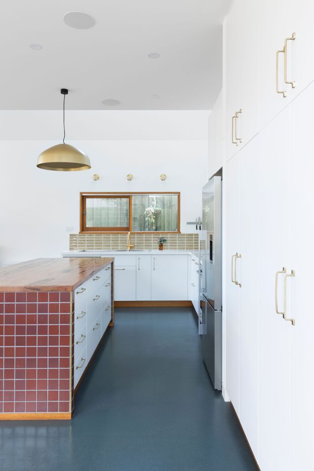 Bright contemporary kitchen with white full-height cabinetry, brass handles, a timber island benchtop, and a red square-tile island face. The kitchen has a dark blue-grey floor, pale tiled splashback, timber-framed window, and a large brass pendant light. The compact layout is clean and strongly colour-blocked with warm timber and brass accents.