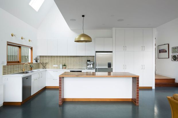 Bright contemporary kitchen with white flat-panel cabinetry, a large central island with a timber benchtop and exposed red-brick side supports, stainless-steel appliances, a muted mosaic-tile splashback, dark flooring, and a brass dome pendant. A skylight and clerestory-like ceiling form bring in natural light, while a timber stair or platform is visible at right.
