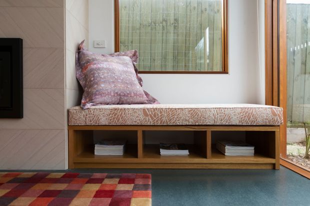 A compact built-in window seat with a patterned upholstered cushion and purple throw pillow, integrated above open timber storage shelving. The seat is beside a large timber-framed window and glazed door, with a colourful geometric rug in the foreground and a dark painted floor. The image has a warm, practical contemporary interior feel with strong timber, patterned textile, and muted jewel-tone accents.