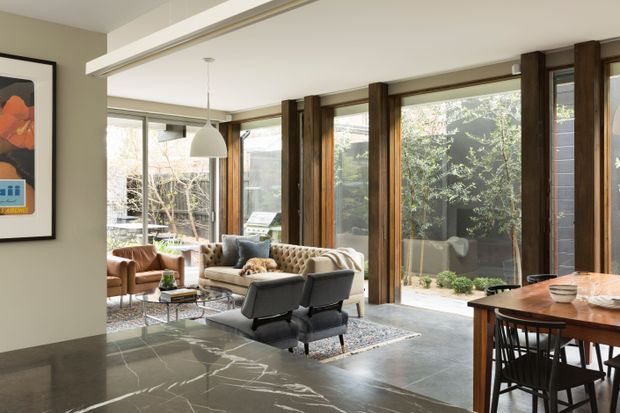 A bright, refined open-plan living and dining space with full-height glazing, dark timber structural framing, polished dark stone flooring, and a warm mix of leather and upholstered seating. The room includes a tufted sofa, lounge chairs, a dining table, pendant lighting, and views toward a landscaped courtyard.