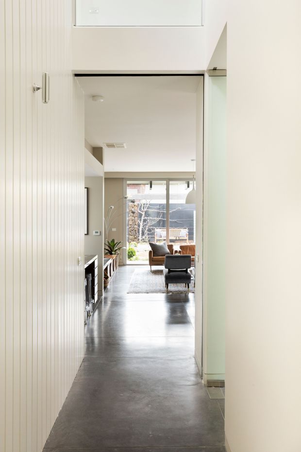 A narrow, bright interior hallway leading toward an open-plan living room. The corridor features vertically panelled white walls, smooth white ceilings, dark polished concrete flooring, minimal built-in cabinetry, and a glazed doorway or opening at the far end. A contemporary living area with lounge seating, a rug, plants, and large glazing is visible beyond.