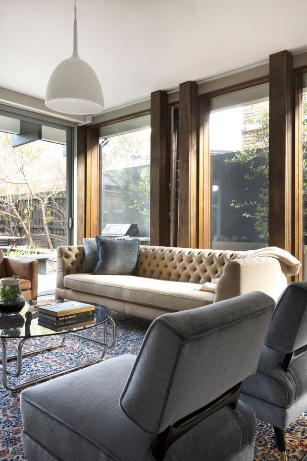 A warmly lit living room with a tufted beige sofa, grey upholstered lounge chairs, a glass coffee table, and a patterned area rug. Full-height timber-framed windows and doors open the room to a leafy courtyard, while a large white pendant light hangs from the ceiling. The room combines contemporary glazing and joinery with comfortable, traditional-inspired furnishings.