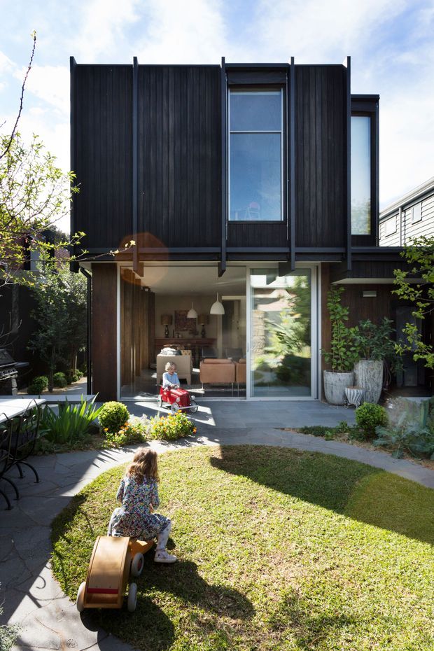 A contemporary multi-level residential extension viewed from a landscaped rear courtyard. The house features dark vertical timber cladding, strong projecting fins, large glazed openings and sliding glass doors, with a warm living area visible inside. A small lawn, paved paths, planting, and children’s toys occupy the foreground garden.