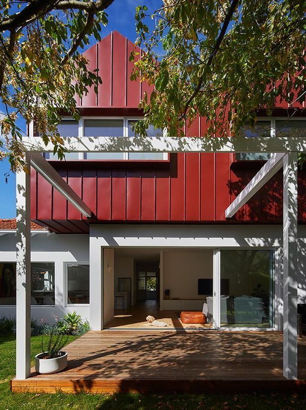 A contemporary two-storey residential extension viewed from a landscaped backyard. The facade combines bold red vertical metal cladding with white rendered walls and a prominent white timber or painted structural frame supporting an upper-level terrace or canopy. Large glazed sliding doors open onto a warm timber deck, with a simple living area visible inside. Mature leafy branches cast patterned shadows across the facade and deck.