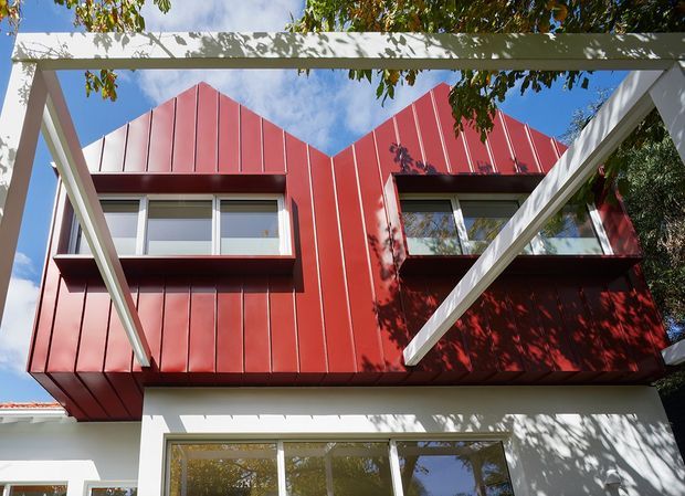 Exterior architectural view of a contemporary multi-level building with a bold red vertically ribbed metal-clad upper facade, angular gabled roof forms, projecting window bays, and white structural framing. Tree shadows fall across the facade, with blue sky visible behind.