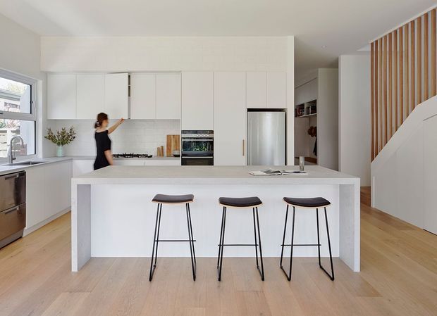Bright contemporary open-plan kitchen with a large white island and three slim black bar stools. The space features white full-height cabinetry, integrated stainless-steel appliances, a pale timber floor, a light stone-like benchtop, and a timber vertical-slat stair screen leading toward the hallway.