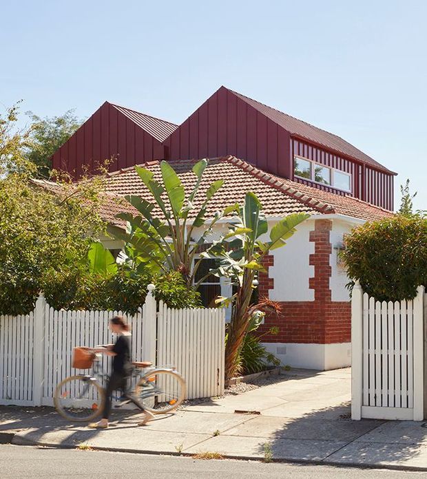 Street-facing view of a renovated residential house with a white-painted brick facade, exposed red-brick detailing, a terracotta tiled lower roof, and a prominent dark red vertical-metal-clad gabled upper extension. A white picket fence, driveway, tropical planting, and a blurred cyclist occupy the foreground.